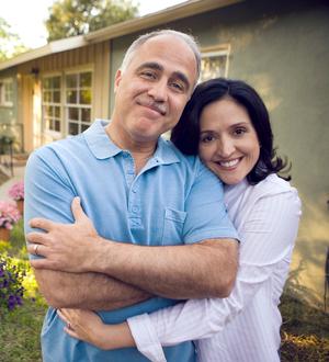 A wife hugs her husband while they both smile for the camera in front of their home.