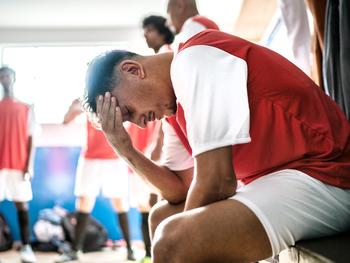 A male soccer player holds his head in pain in the locker room after a game.