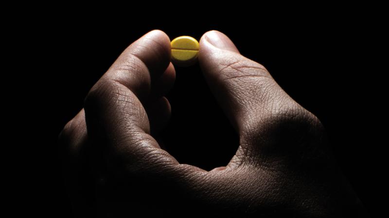 Close up photo of a hand holding a single pill, with dramatic lighting.