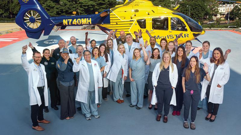 A multi-disciplinary team of healthcare professionals poses for a group photo in front of the MedStar Helicopter at MedStar Washington Hospital Center.