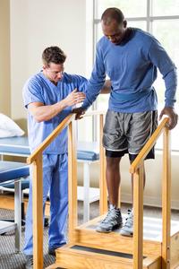 A therapist helps a male patient walk down stairs in a rehabilitation gym.