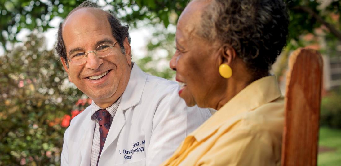 Dr. David Shocket, gastroenterologist, sits and talks with a female patient. Both are sitting together on a bench in a garden.