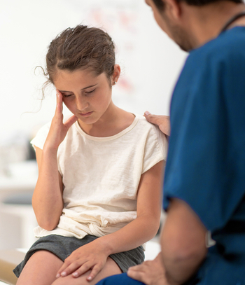 A young girl holds her head in pain while talking to a doctor.