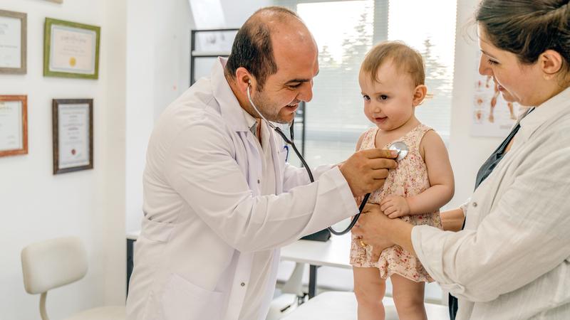 A doctor listens to the heart of a small child while a nurse holds her, during a well-care visit.