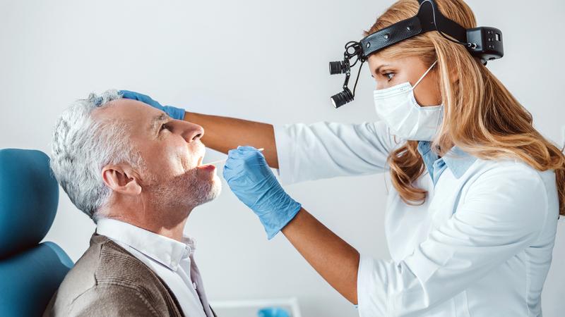 A doctor examines the mouth of an older male patient using magnifying lenses.