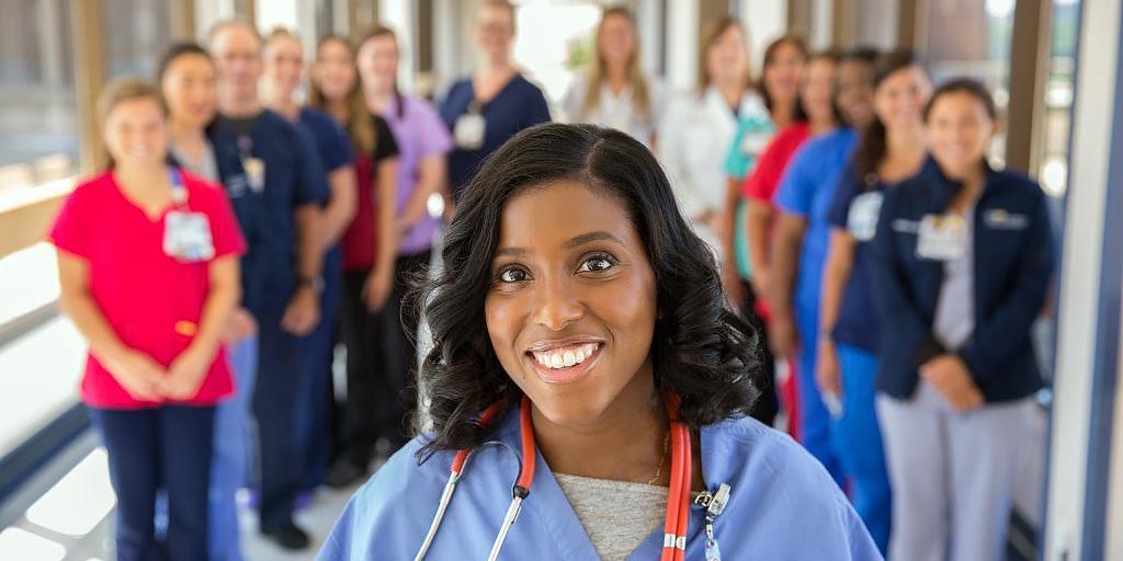 A nurse poses for a photo in a hospital hallway, while a group of fellow nurses stands in the background.