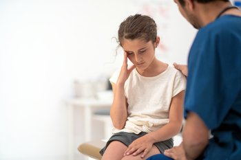 A young girl holds her head in pain while talking to a doctor.