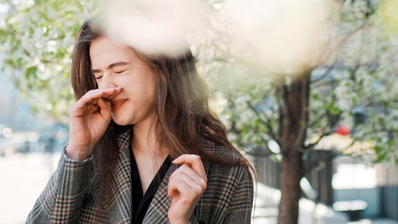 A professional woman has an itchy nose standing outside in the springtime.