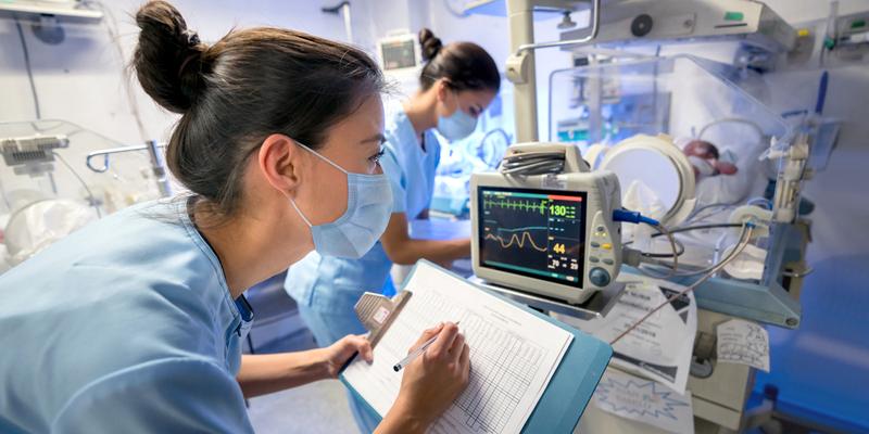 Two healthcare professionals tend to a patient, and record data from a machine monitoring vital signs.