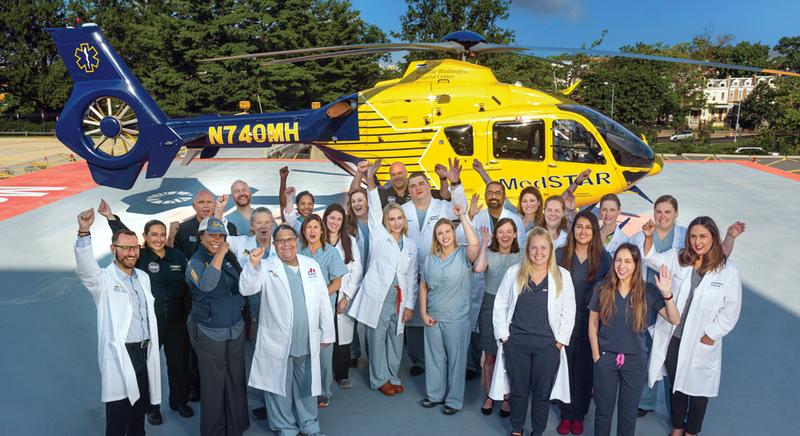 A multi-disciplinary team of healthcare professionals poses for a group photo in front of the MedStar Helicopter at MedStar Washington Hospital Center.