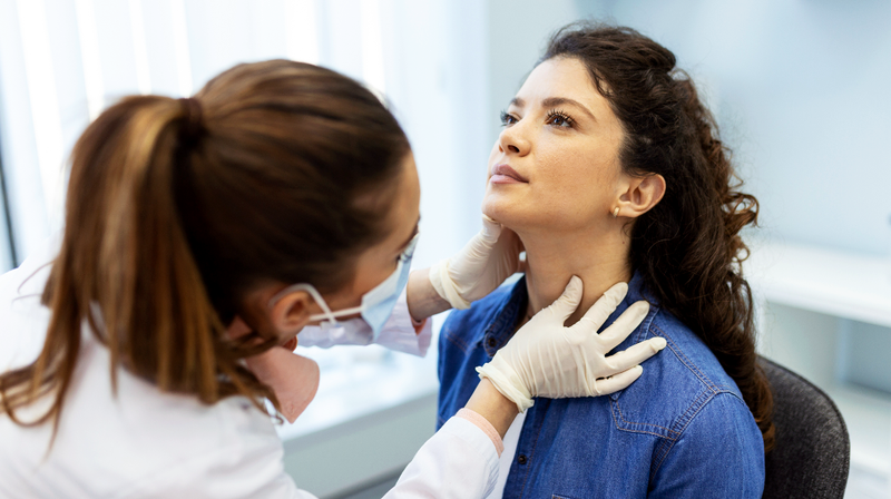 A female doctor examines the neck of a female patient in a clinical setting.