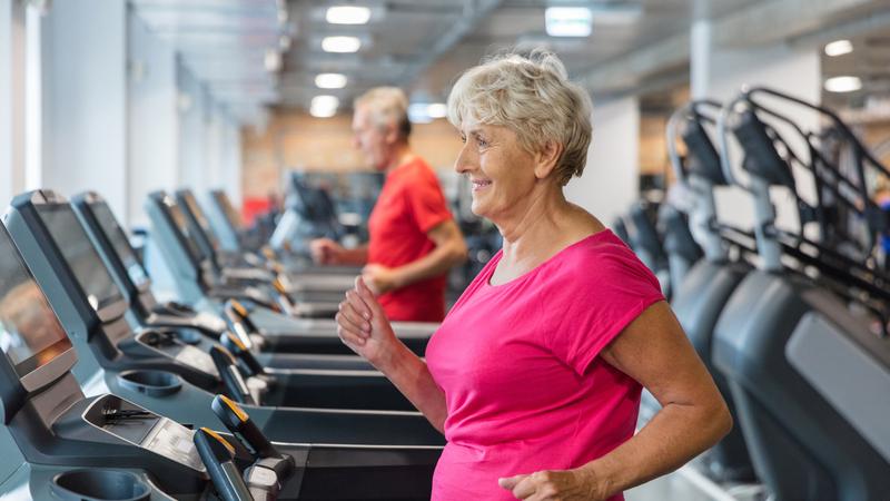 A senior woman wearing a bright pink shirt smiles while walking on a treadmill in a gym.