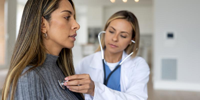 A female doctor listens to the heart of a young female patient in a clinical setting.