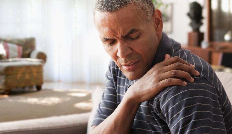 A mature African-American man holds his shoulder while wincing in pain. He is sitting on a sofa in his livingroom.