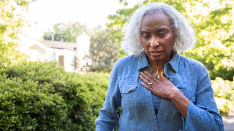 A woman shows a look of concern as she puts her hand on her chest in discomfort while standing outdoors.