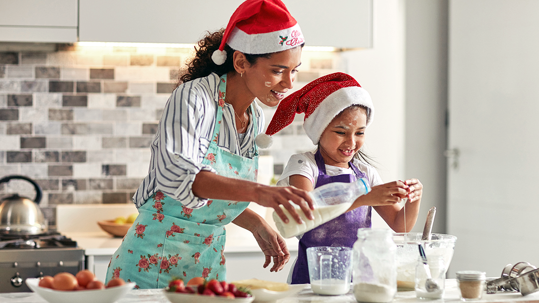 A mother and her young daughter prepare food in the kitchen while wearing Santa hats.