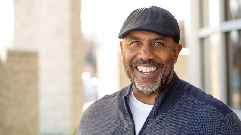 A handsome middle aged man wearing a hat smiles for the camera.