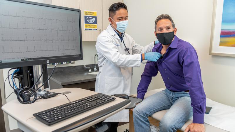 A doctor listens to a patient's heart during an office visit. Both people are wearing masks.