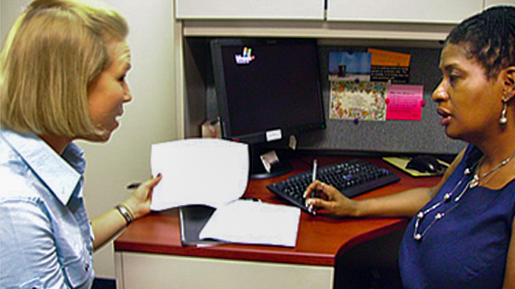 Two women sit together in an office cubicle and talk.