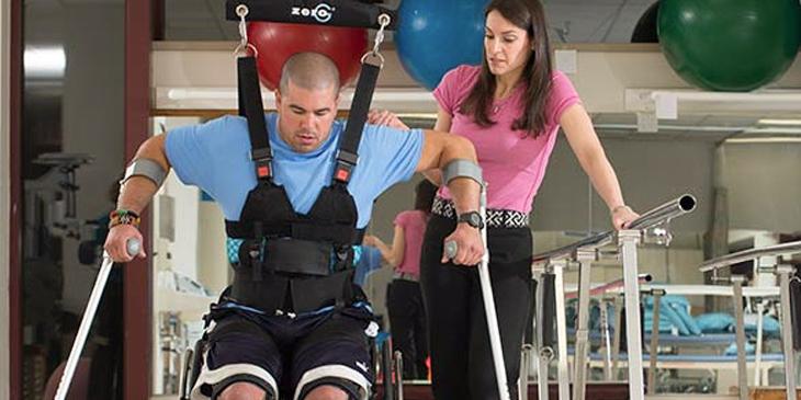 在MedStar国家康复医院的康复健身房，一名理疗师正在帮助一名脊髓/脑损伤患者学习使用拐杖和腿部支架走路。