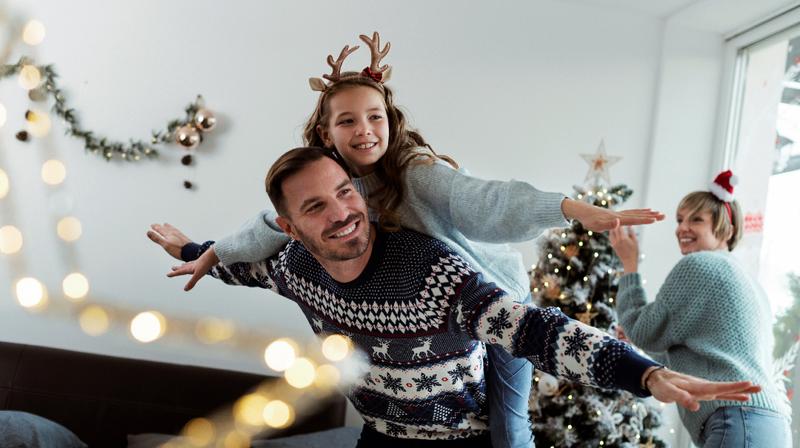 A father carries his daughter on his back in the living room of his home decorated for Christmas.