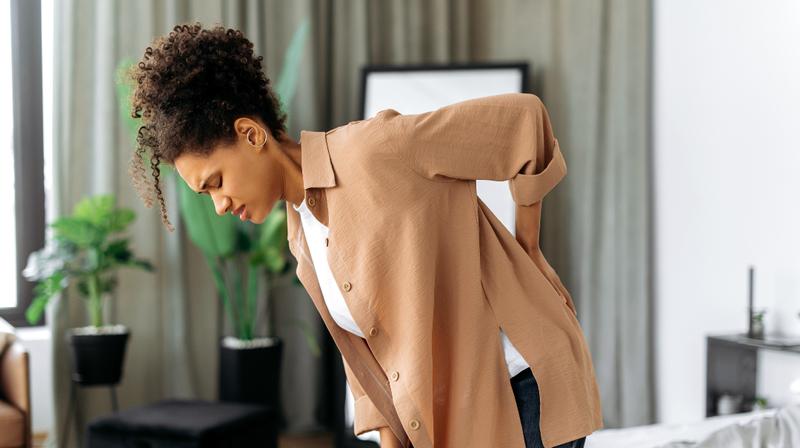 A woman bends over and puts her hand on her lower back because she is in pain.