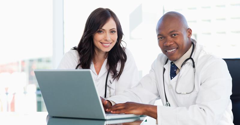 Two doctors, a man and a woman, working in front of a grey laptop while sitting at the desk