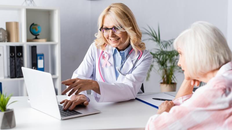 A female provider, wearing a lab coat and a pink stethoscope around her neck, consults with a mature female patient in an office setting.