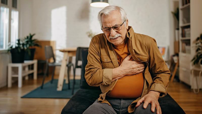 An older man with grey hair sits in a chair in his home and clutches his chest while he winces in pain.