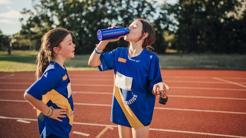 2 young girls drink water after running on an outdoor track.
