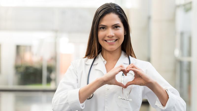 A young female medical professional wearing a white lab coat stands in a hospital hallway and makes a heart symbol with her hands. She is smiling and looking at the camera.