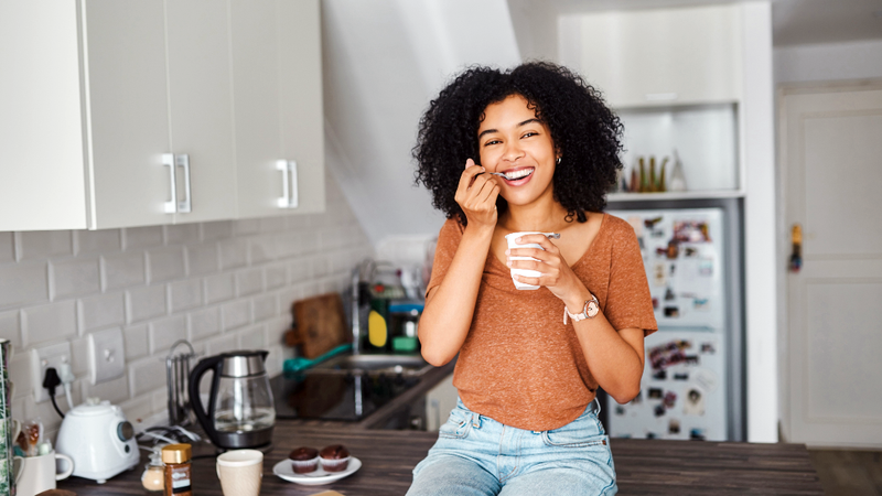 A young woman sits on her kitchen counter and eats healthy food.