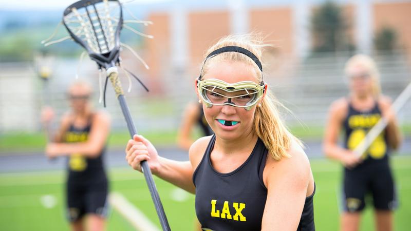 A young woman dressed to play lacrosse holds her stick and smiles for the camera. She is wearing a mouth guard and eye protection, and her teammates are out of focus in the background.