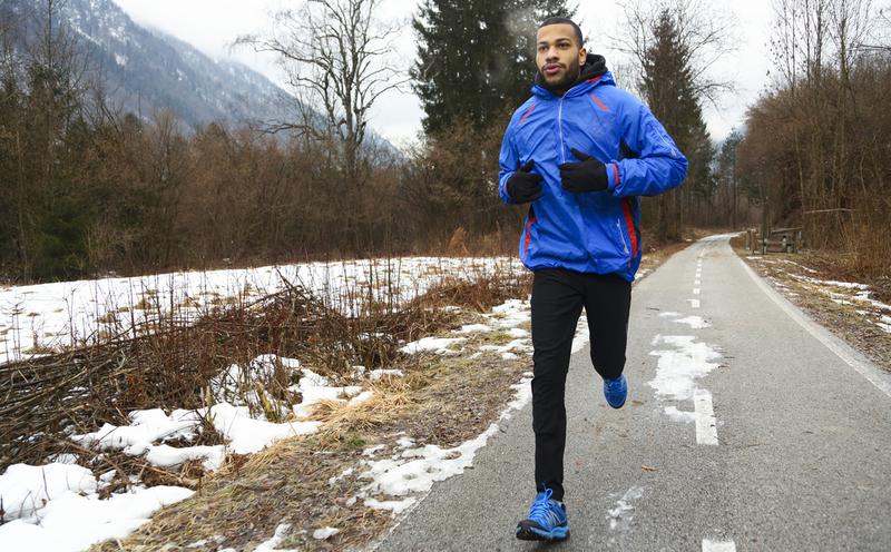 A young african american man in sports clothing running on a rural mountain trail road in winter. Snow on road and fog in the park.