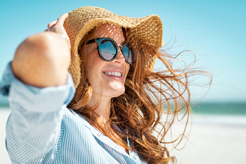 Side view of a beautiful mature woman wearing sunglasses enjoying at beach. Young smiling woman on vacation looking away while enjoying sea breeze wearing straw hat. Closeup portrait of a girl relaxing at sea.