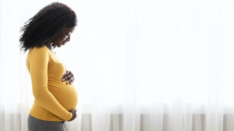 A pregnant woman, wearing a yellow sweater, looks down at her belly as she stands in front of a window.