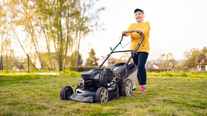 A woman wearing a yellow shirt mows her lawn with a push mower.