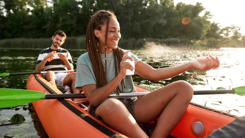 A young couple sits in an orange kayak on a lake. The young woman is applying sunscreen spray.