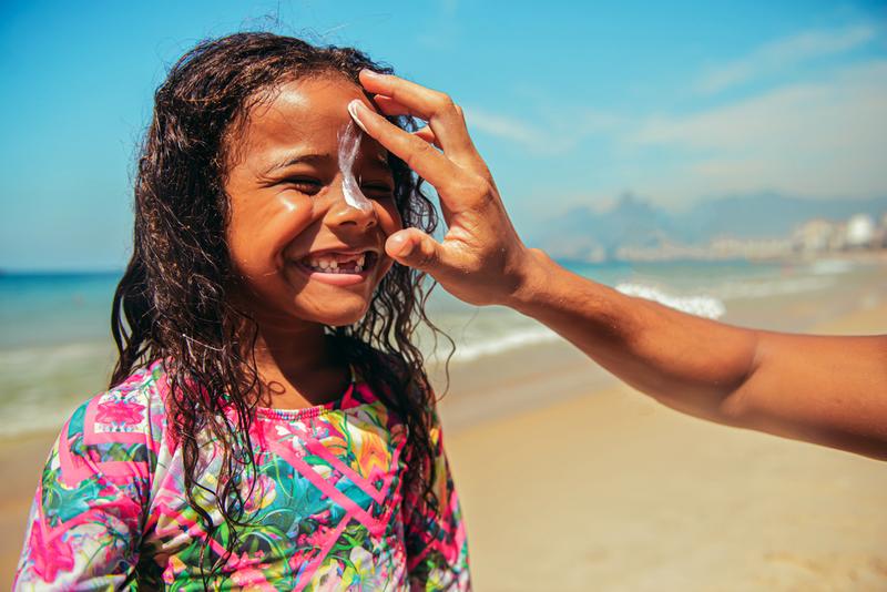 Close up photo of a smiling little surfer girl who is having an adult put sunscreen lotion on her nose.