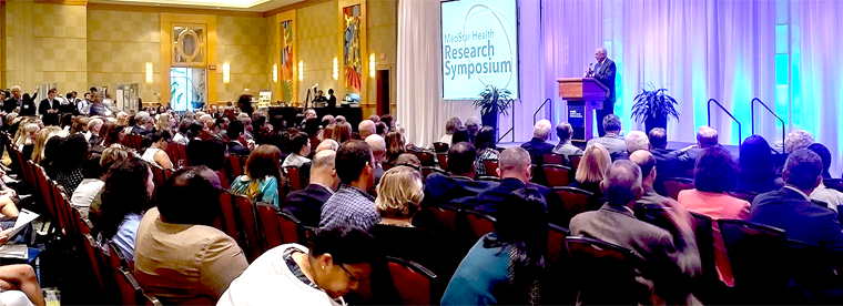 A speaker stands at a podium in front of a large audience in an auditorium at a professional symposium event.