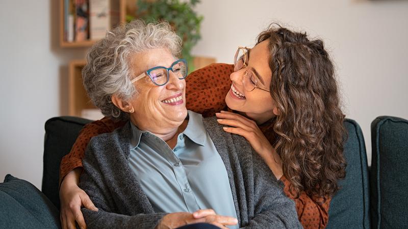 A young woman smiles at her older female family member sitting in an easy chair in her home.