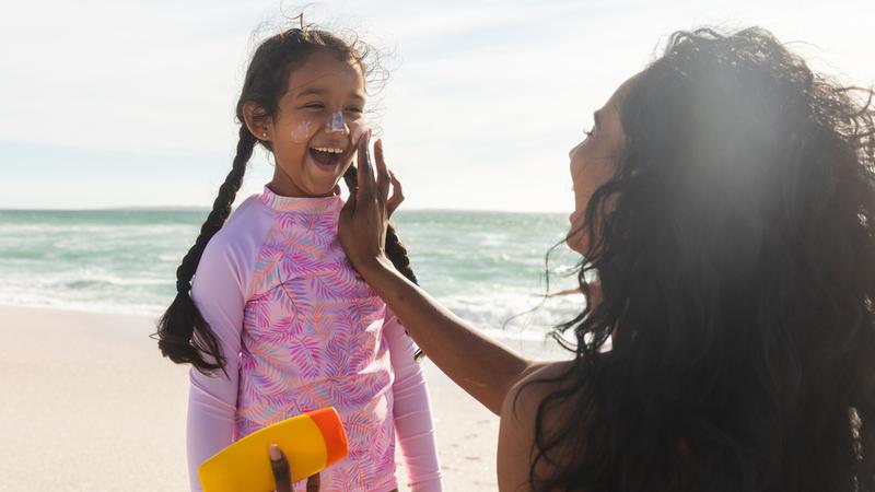 A smiling little surfer girl who is having her mother put sunscreen lotion on her nose.