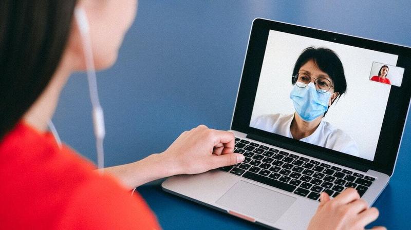 A young woman looks at a laptop computer to have a video telehealth visit with a doctor. The doctor is wearing a mask.