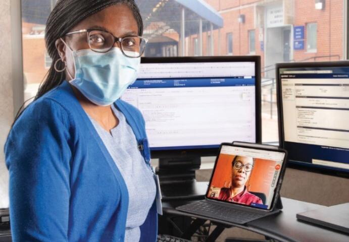 A MedStar Health provider poses for a photo in front of several computers, one of them with a female patient who has called in for a telehealth visit.