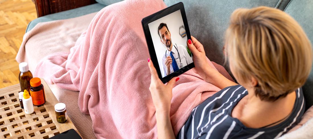 A woman sits on her sofa under a pink blanket and talks with a doctor during a telehealth appointment.