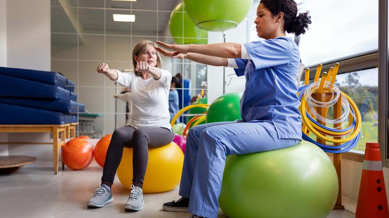 A female therapist wearing blue scrubs sits on a yoga ball with her arms outstretched while her female patient mirrors her during a physical therapy session in a rehabilitation gym.