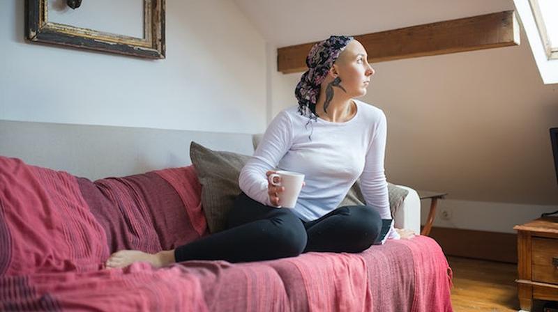 A young woman undergoing treatment for cancer sits on her sofa and looks out the window.