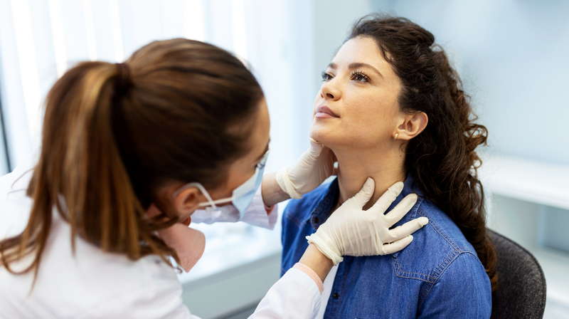 A female doctor examines the neck of a female patient in a clinical setting.