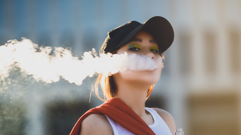 A young woman blows a cloud of smoke while vaping.