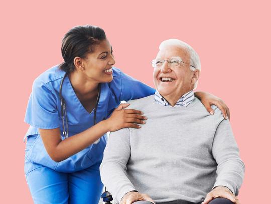 A female healthcare provider wearing blue scrubs shares a laugh with a mature adult male patient.